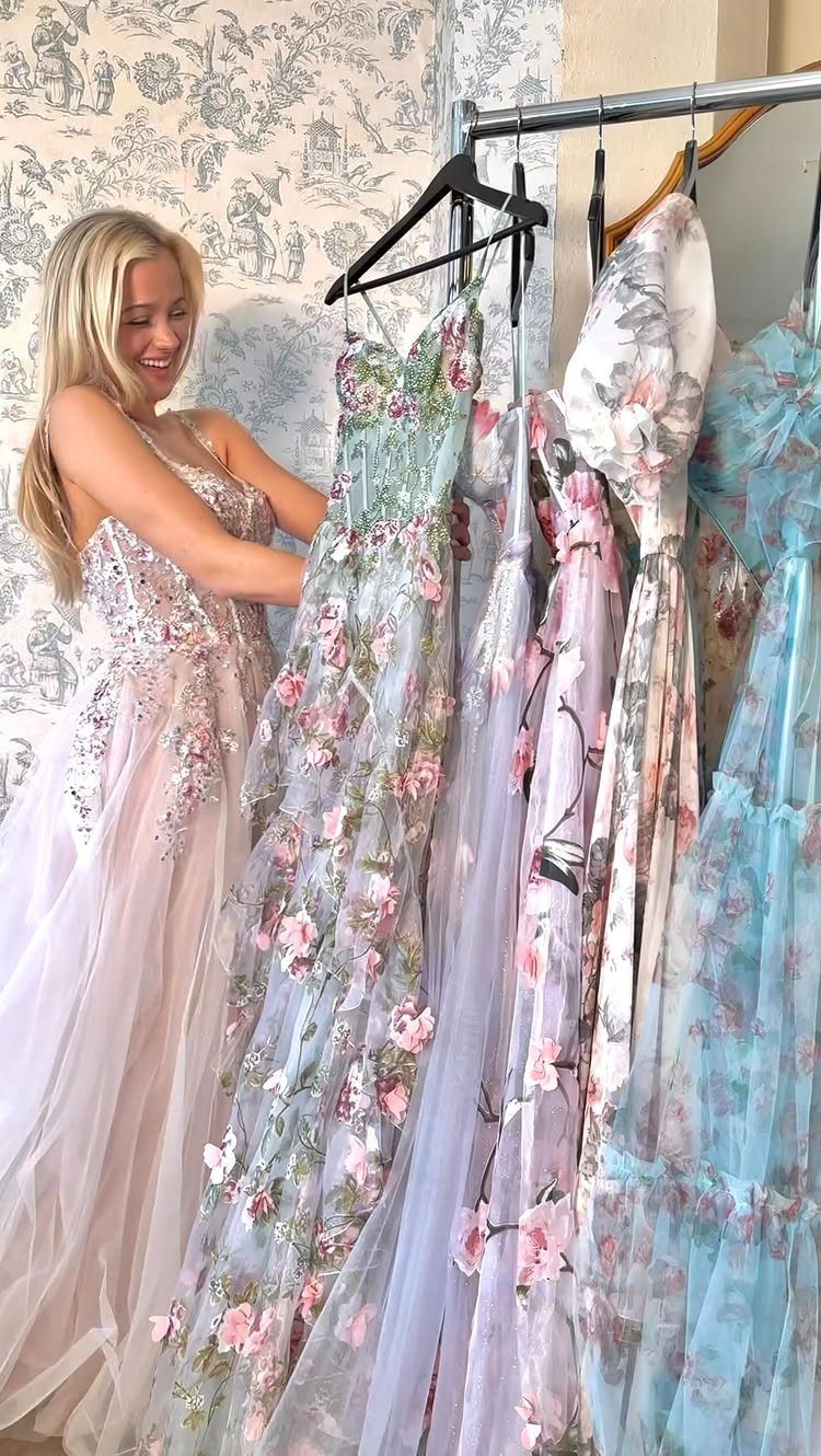 10 Nice Prom Dresses for Every Style and Body Type 7 Woman in pink gown standing beside a rack of floral prom dresses.