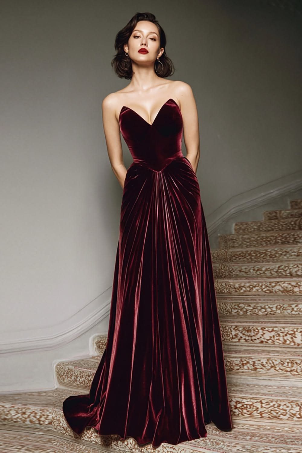 Woman in a strapless burgundy velvet gown with a pleated skirt on stairs.