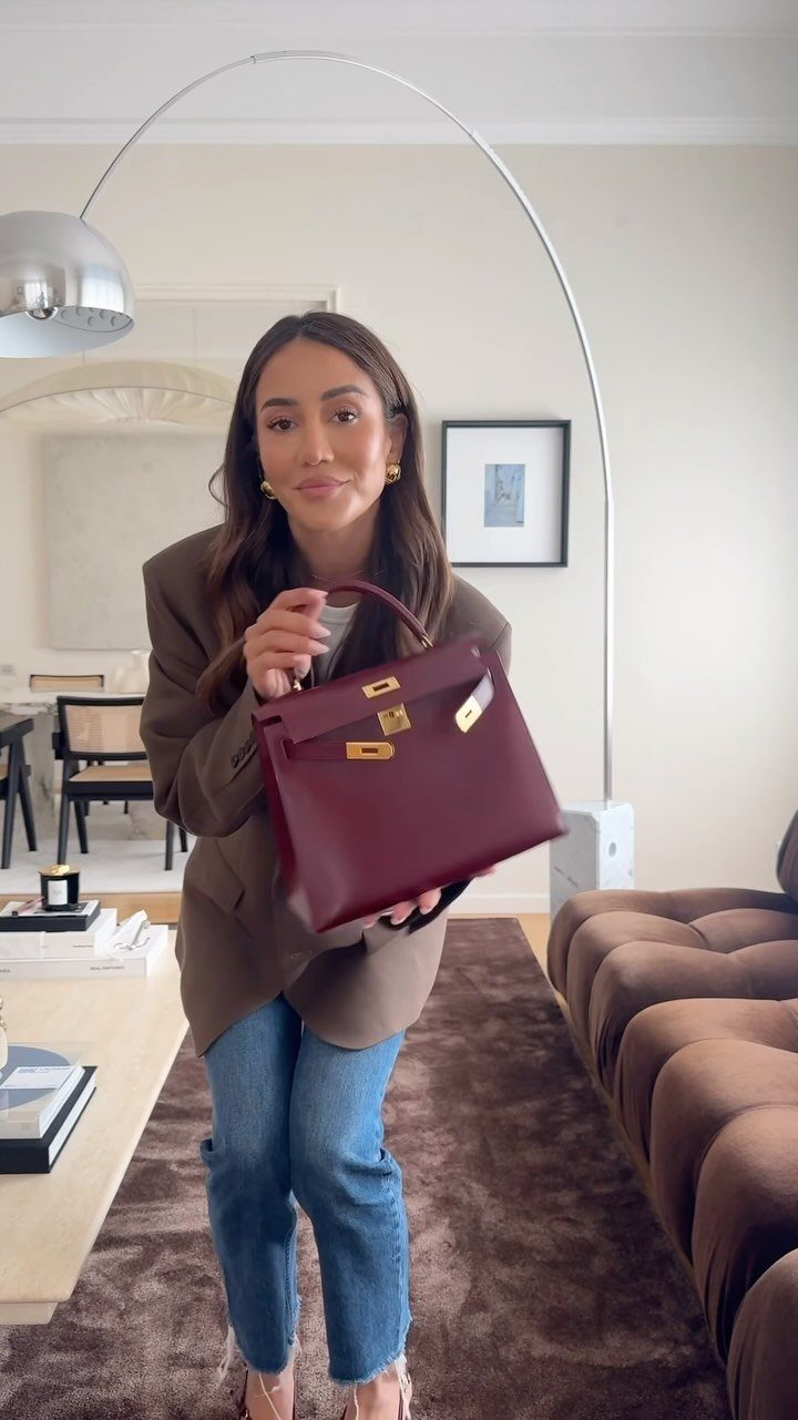 Woman wearing a brown blazer and blue jeans holding a burgundy leather handbag.