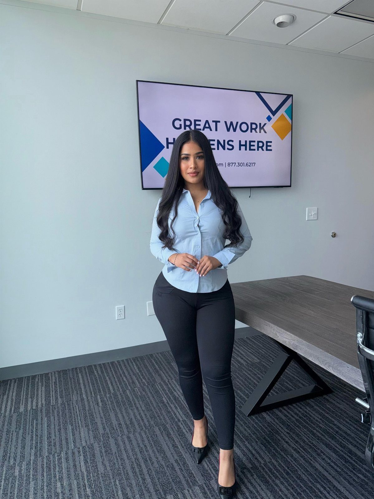 Woman in light blue button-down shirt and black skinny jeans in an office.
