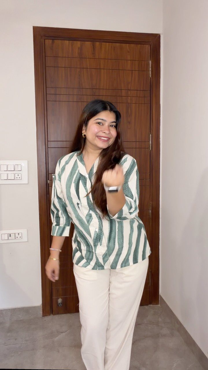 Woman wearing a green and white printed shirt with white trousers standing indoors.