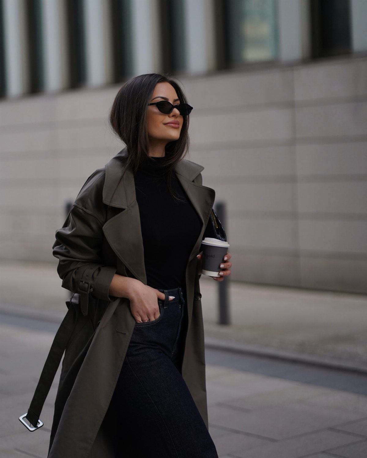 Woman in olive green trench coat, black turtleneck, and dark blue jeans holding coffee.
