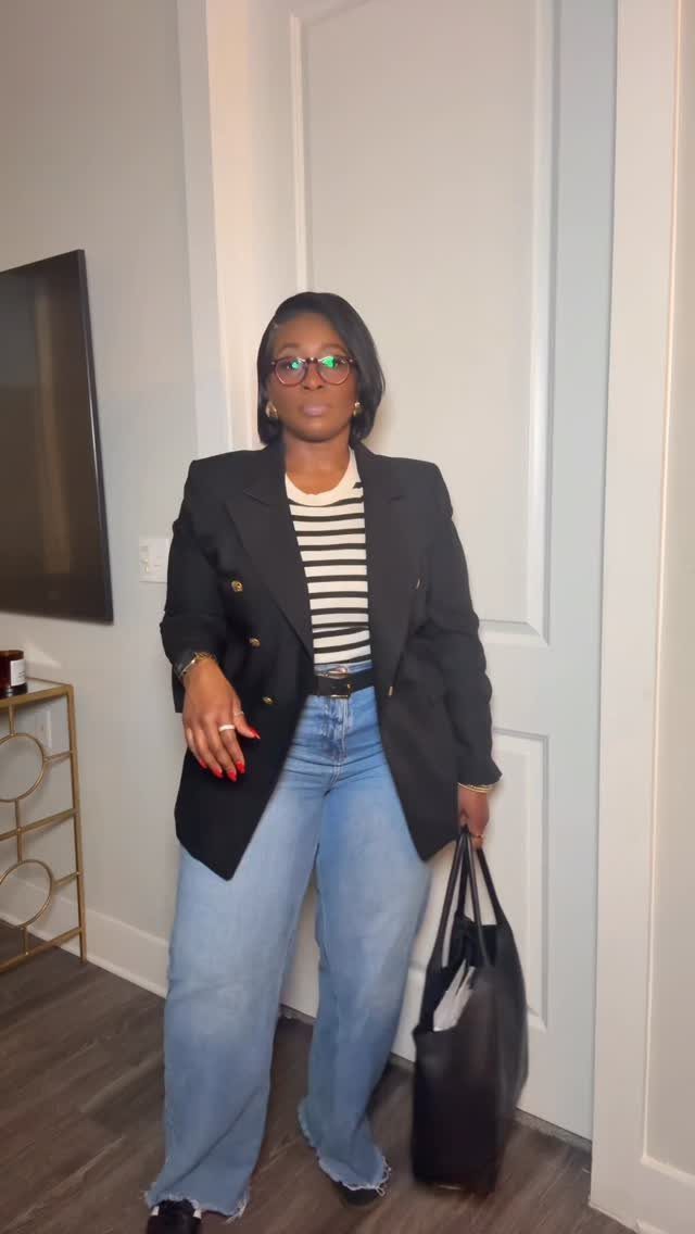 Woman in black blazer and wide-leg blue jeans holding a black tote bag.