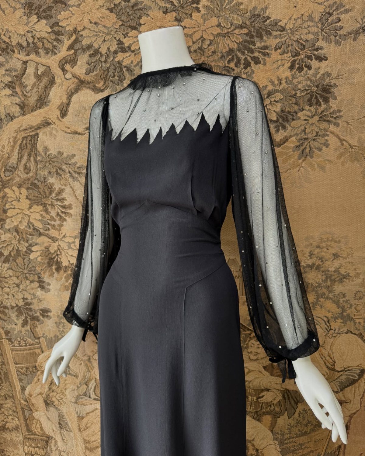 Old Fashion Designers and 3 Ideas They Made Classic 1 Black vintage dress with sheer beaded sleeves on a white mannequin against tapestry