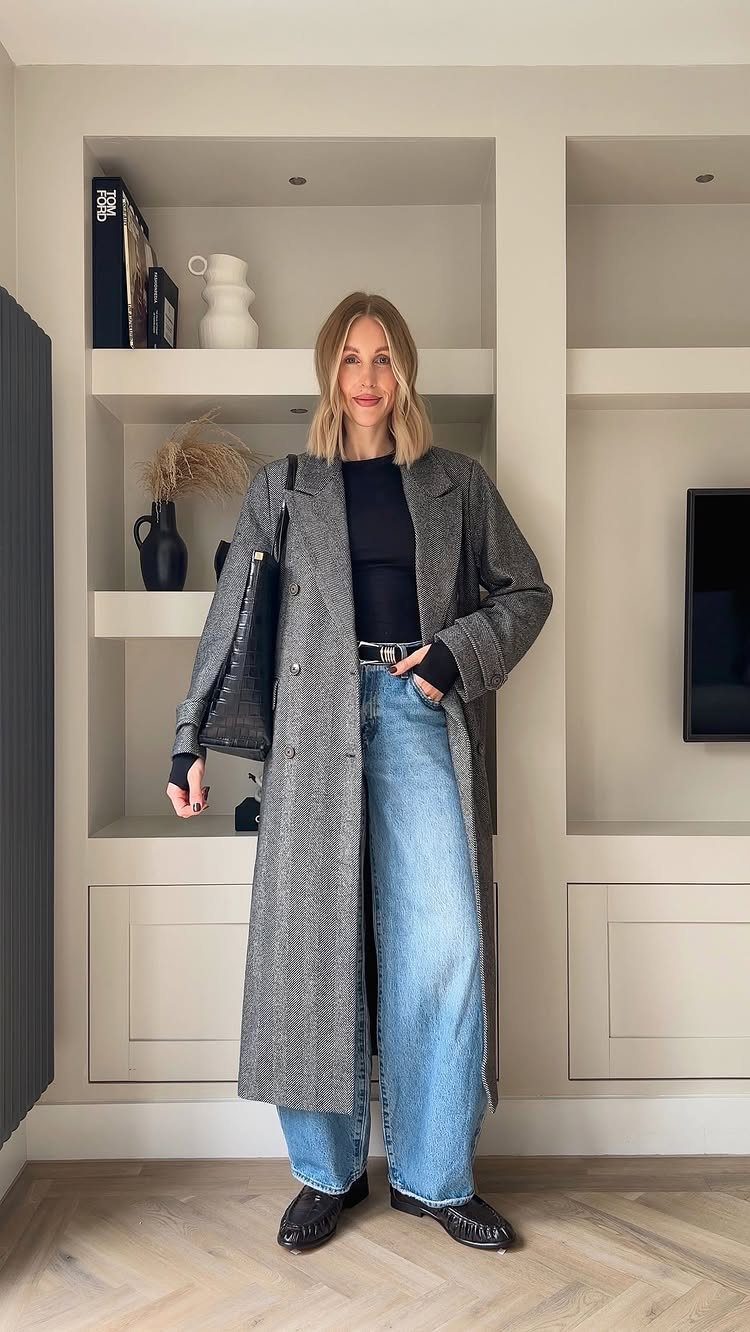 Woman wearing a long grey coat, black top, and wide-leg blue jeans indoors.