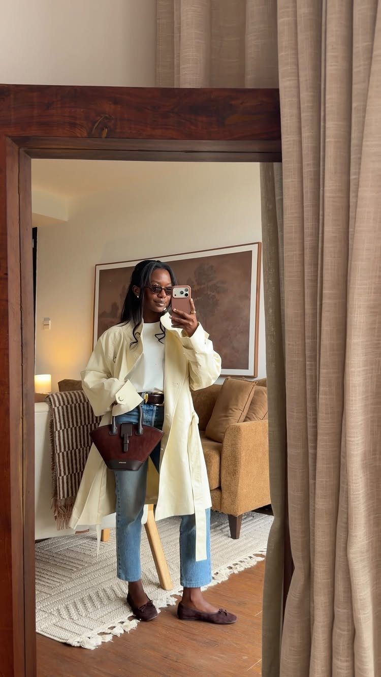 Woman in cream coat and blue jeans wearing dark brown ballet flats.