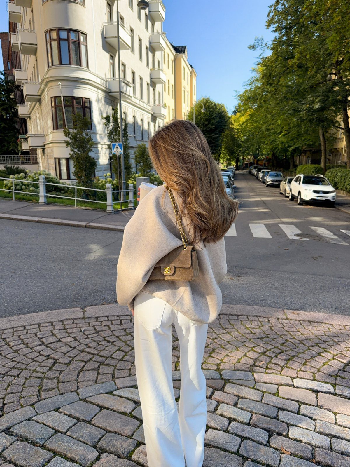 Woman in beige sweater and white pants standing on a cobblestone city street.