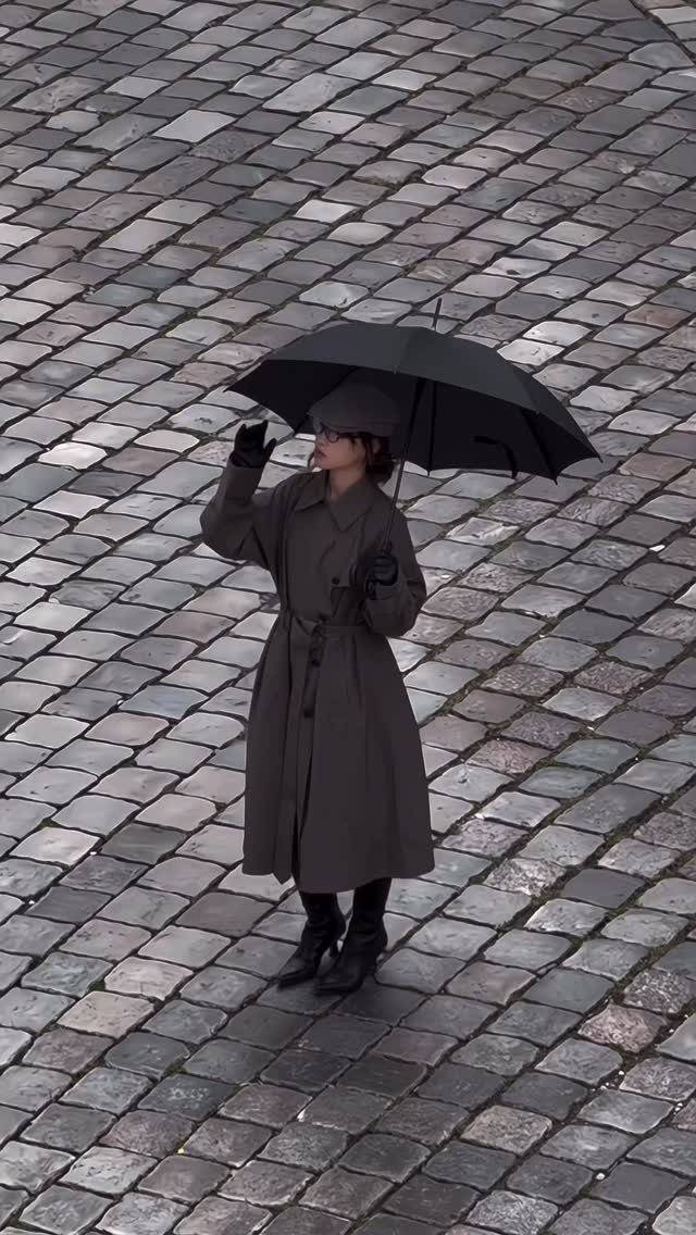 Person in a taupe trench coat and beret holding a black umbrella on cobblestones