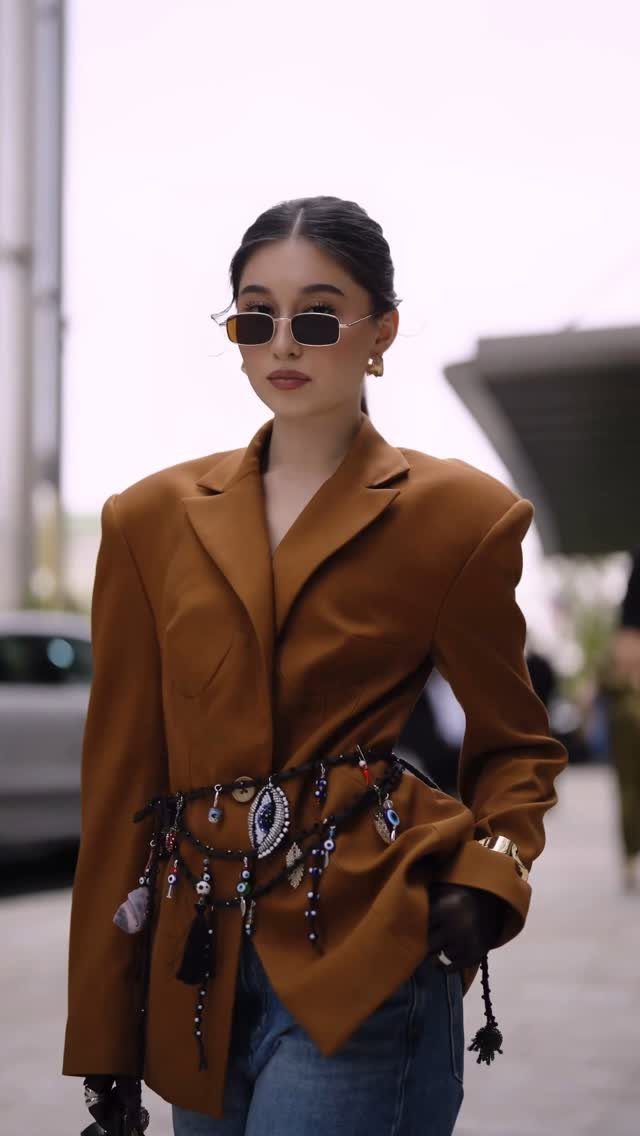 Woman in a brown structured blazer and beaded belt walking outdoors in sunglasses.