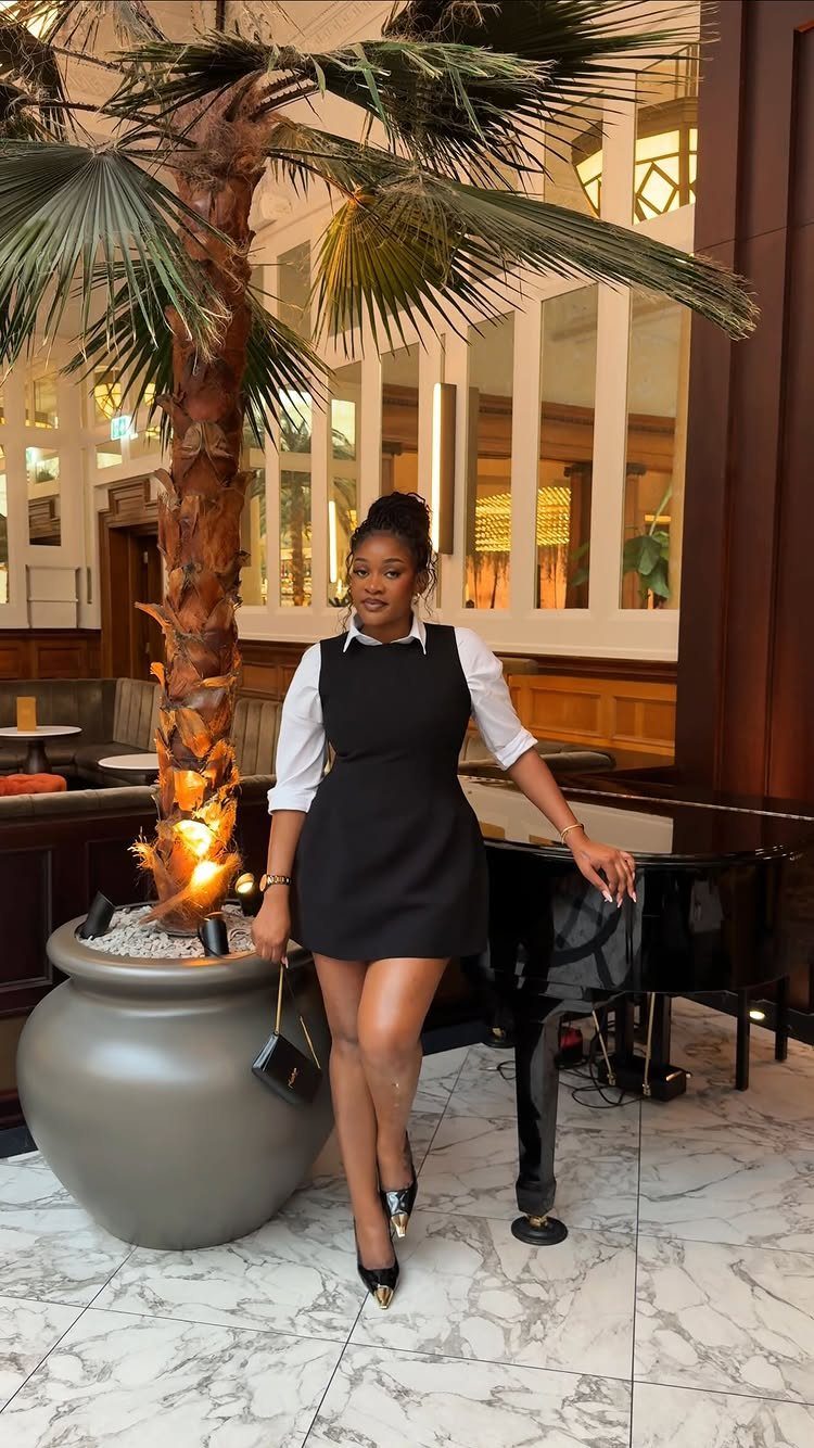 Woman in a short black dress over a white shirt standing by a piano.