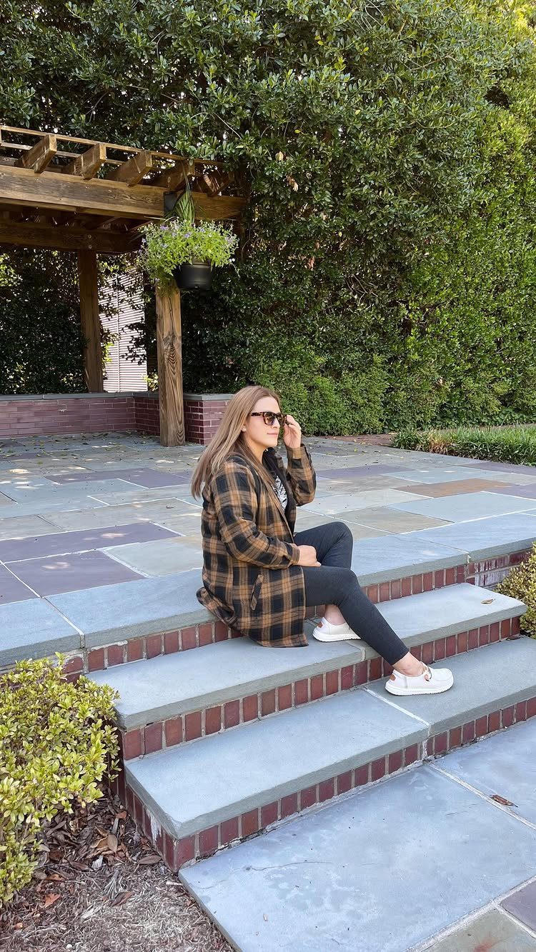 Woman in brown plaid shirt and black skinny jeans sitting on stone steps.