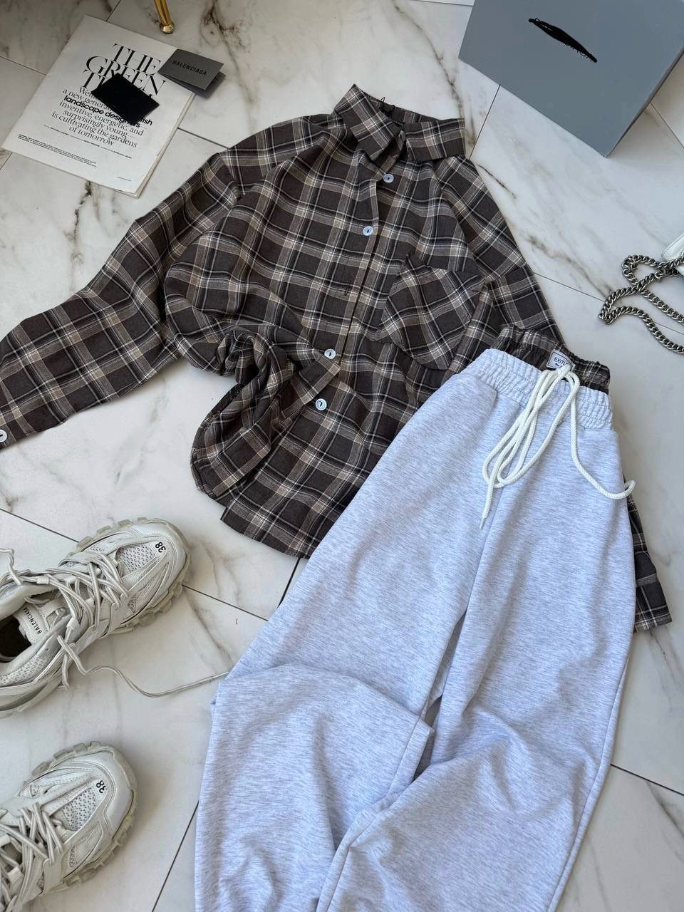 Brown plaid shirt, light grey joggers, and white sneakers arranged on marble surface.