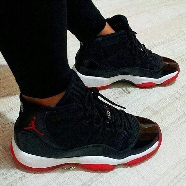 Black Air Jordan 11 sneakers with red soles worn with black pants.