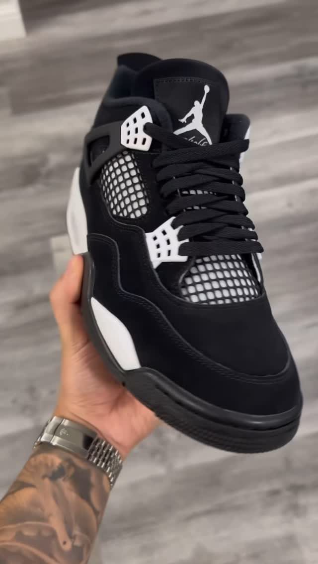 Black Air Jordan 4 sneaker with white accents held by a tattooed hand.
