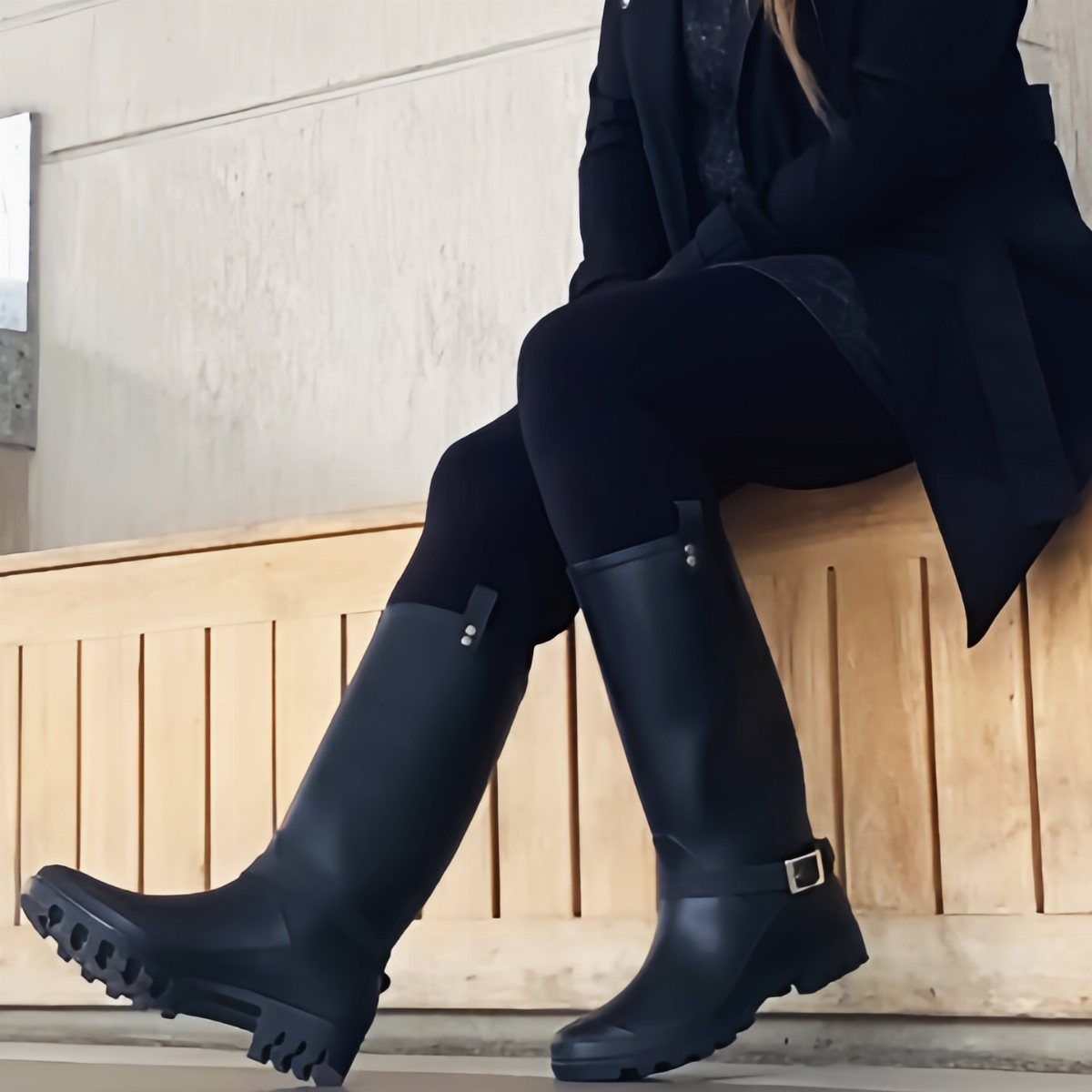 Black rubber rain boots with lug soles worn by a person on a bench.
