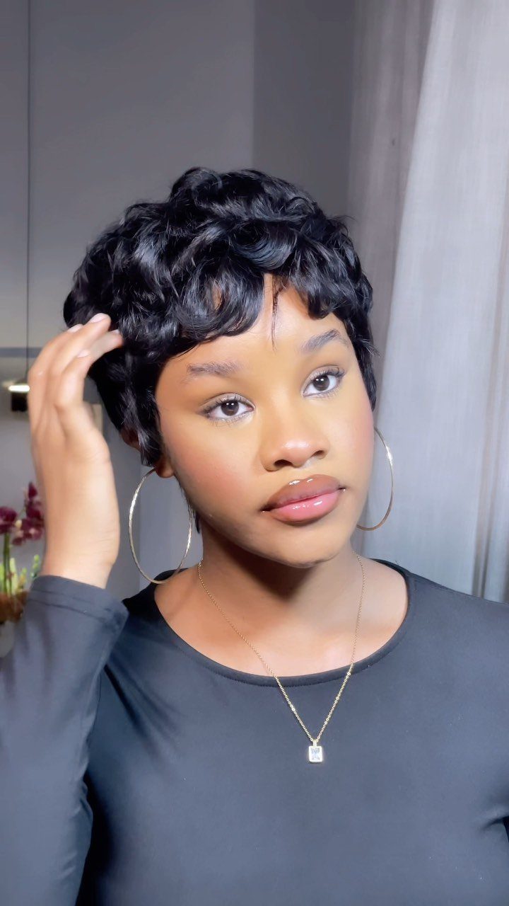 Woman wearing a short curly black pixie cut wig and gold jewelry.