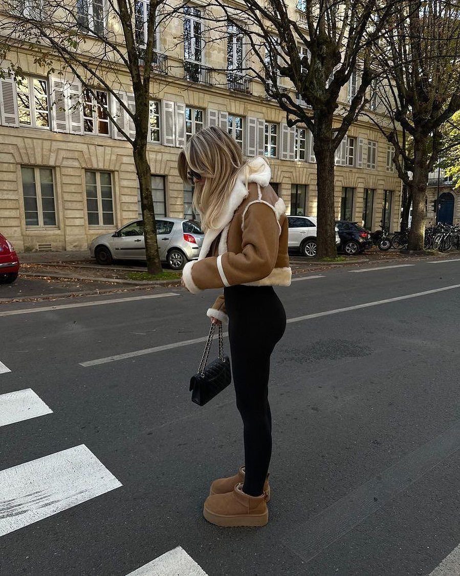 Short UGG Boots: 7 Outfit Ideas for Jeans, Skirts and Blazers 3 Woman wearing chestnut short UGG boots, black leggings, and a tan shearling jacket.
