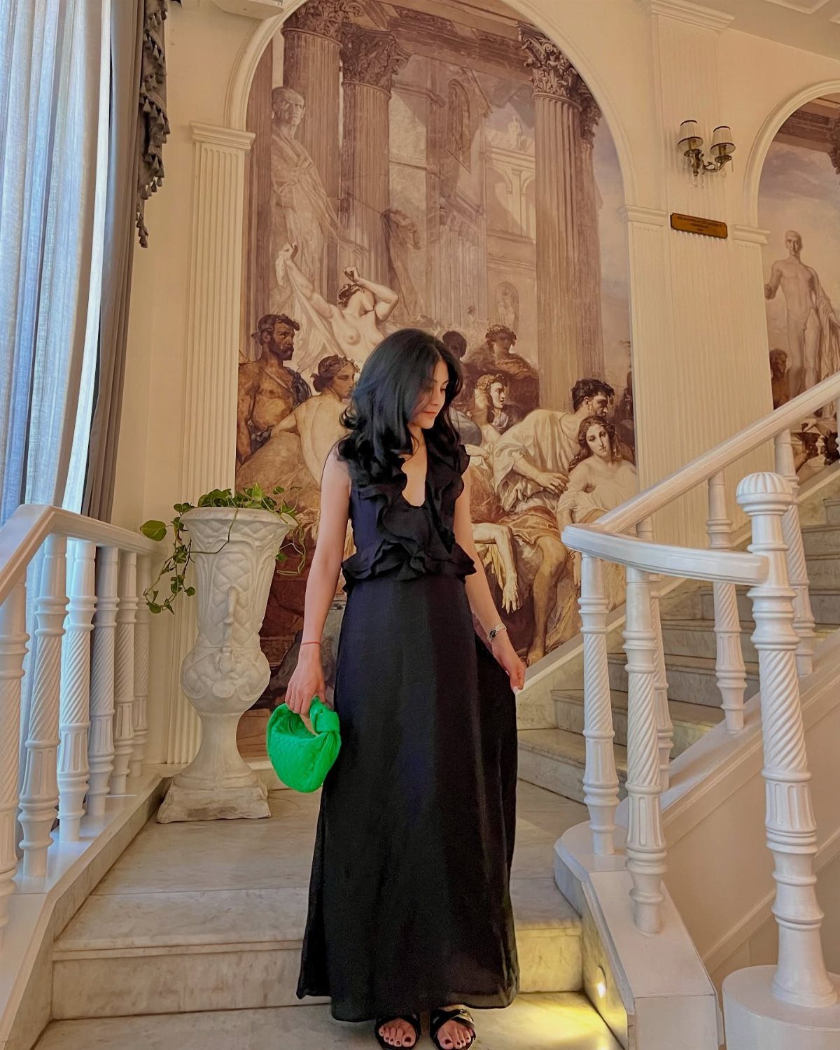 Woman in a black ruffle maxi dress holding a green bag on marble stairs