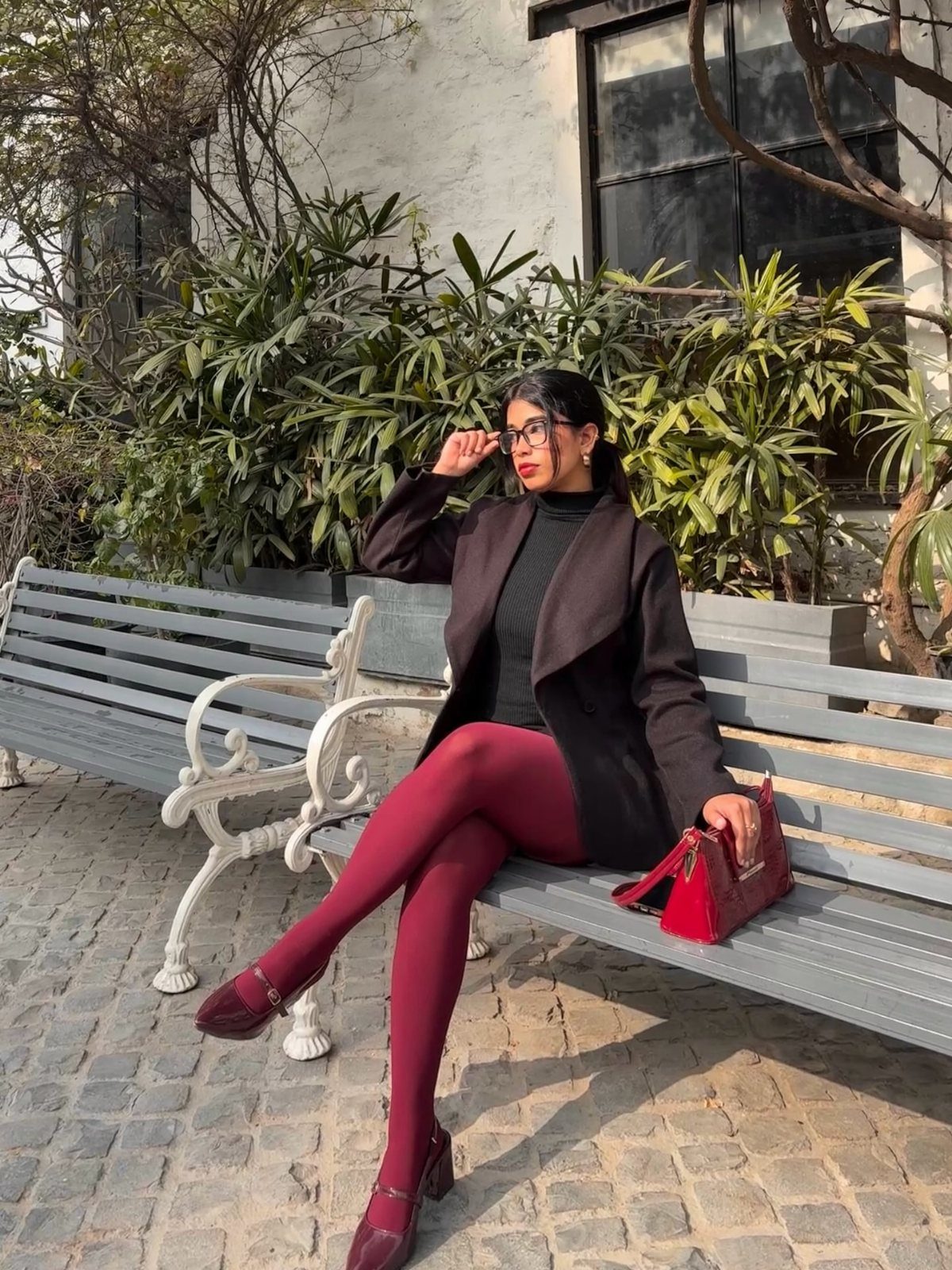 Woman in dark blazer and burgundy tights sitting on a grey park bench.