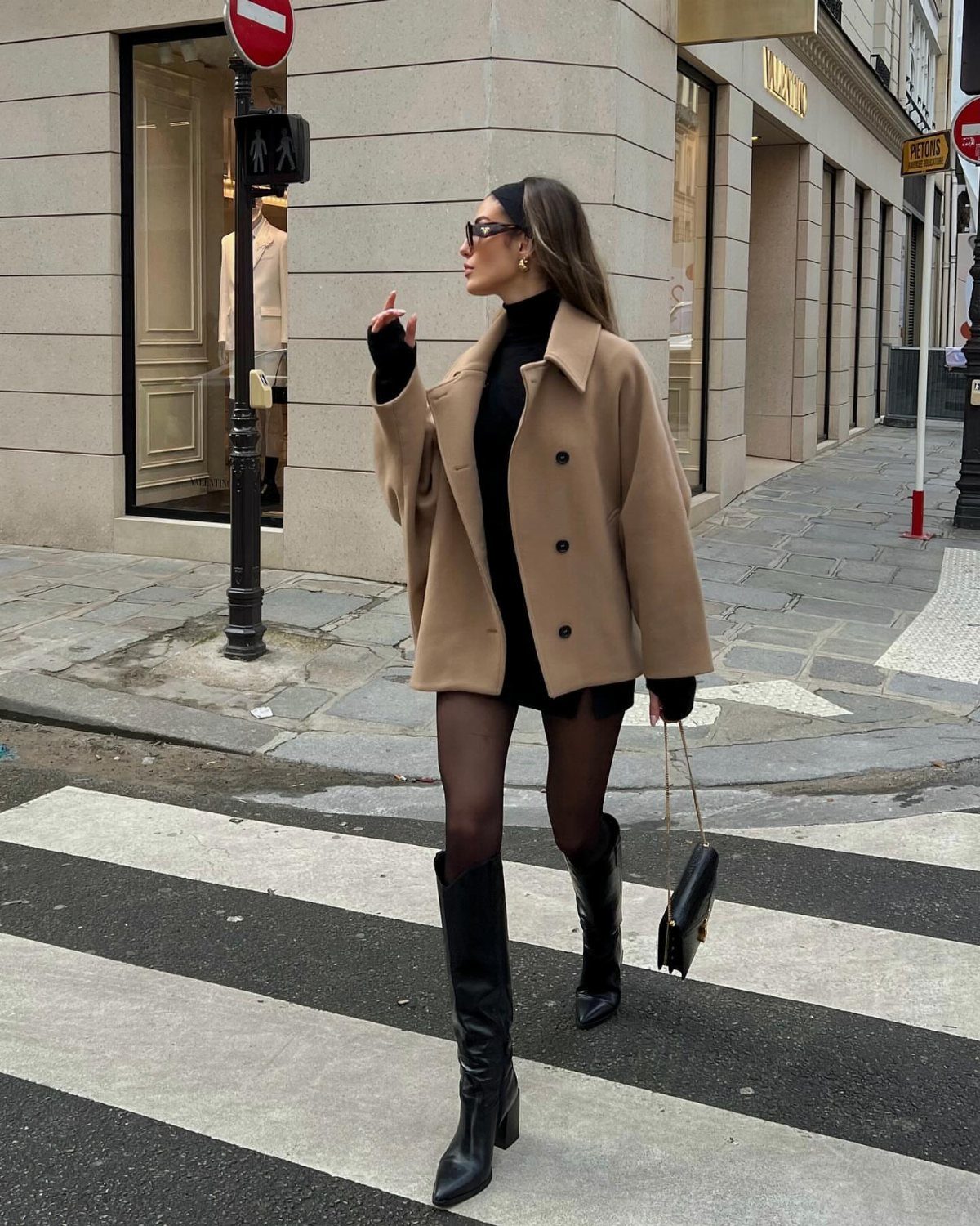 Woman in camel coat, black mini dress, and sheer black tights crossing street.