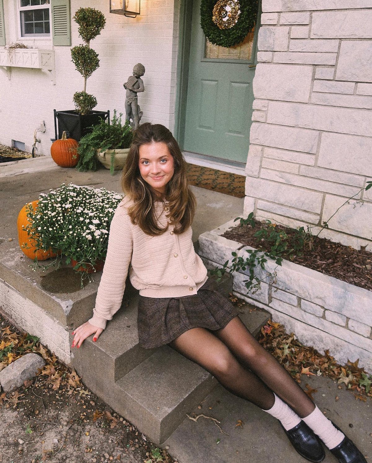 Woman in beige cardigan, plaid skirt, and black tights sitting on porch steps.