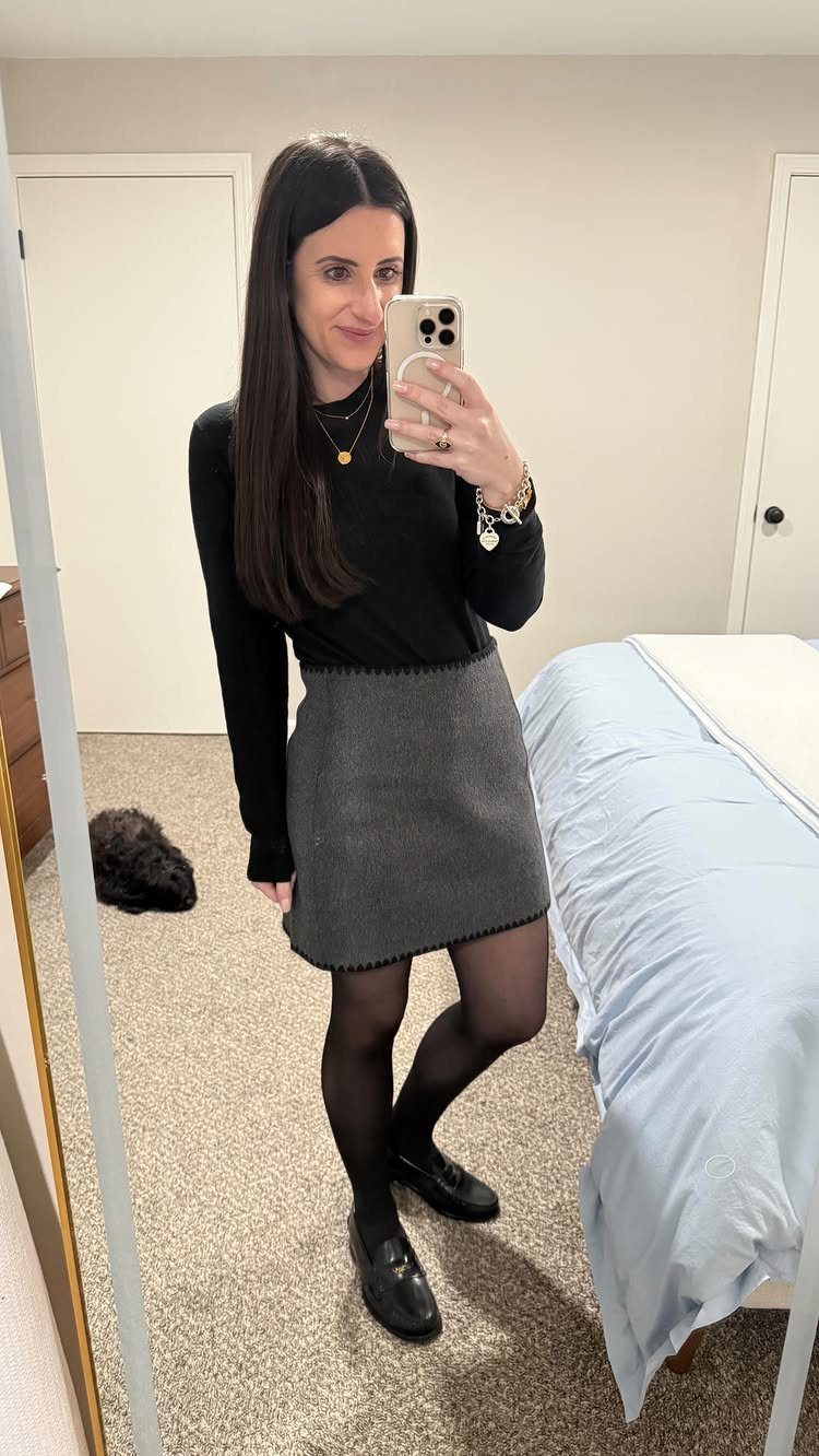 Woman wearing a black top, grey mini skirt, and black tights in bedroom