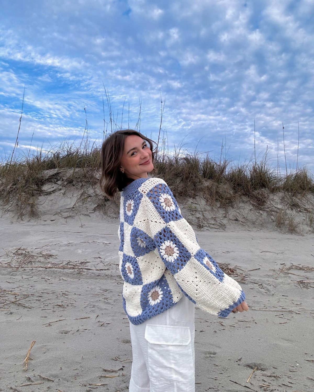 Woman on a beach wearing a blue and white crochet floral cardigan and white pants