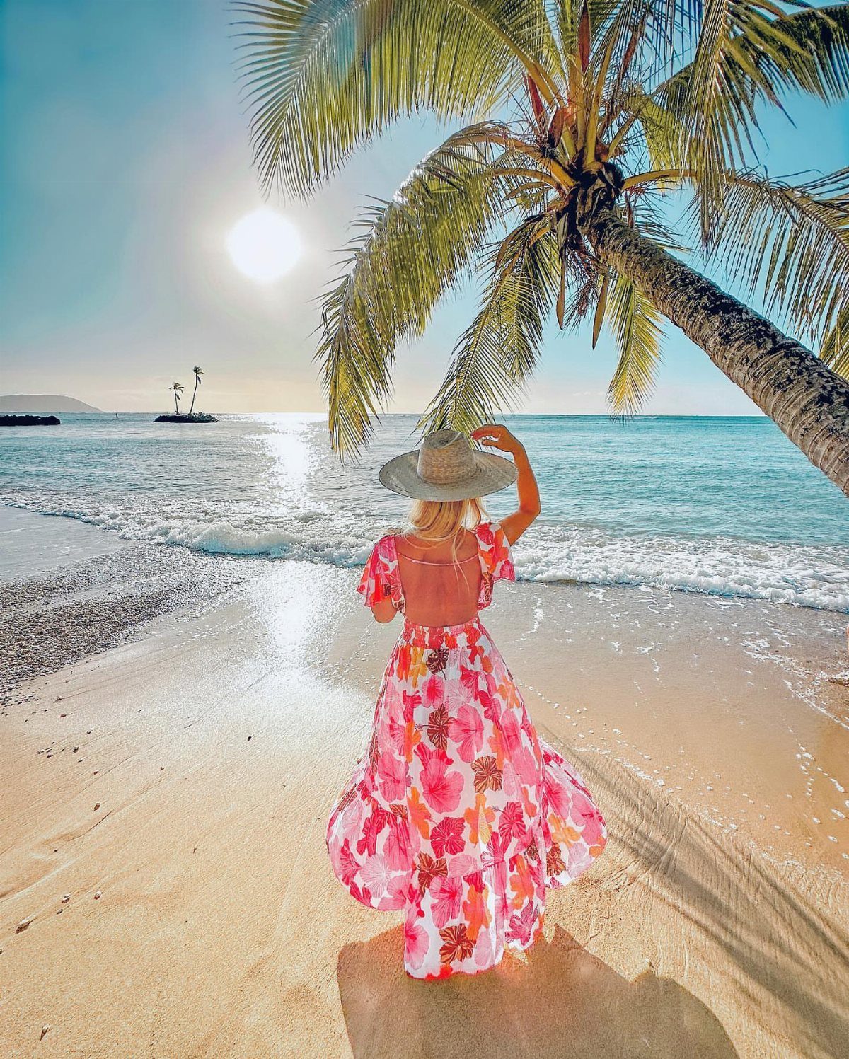 Woman in a floral maxi dress and straw hat standing on a sunny beach.
