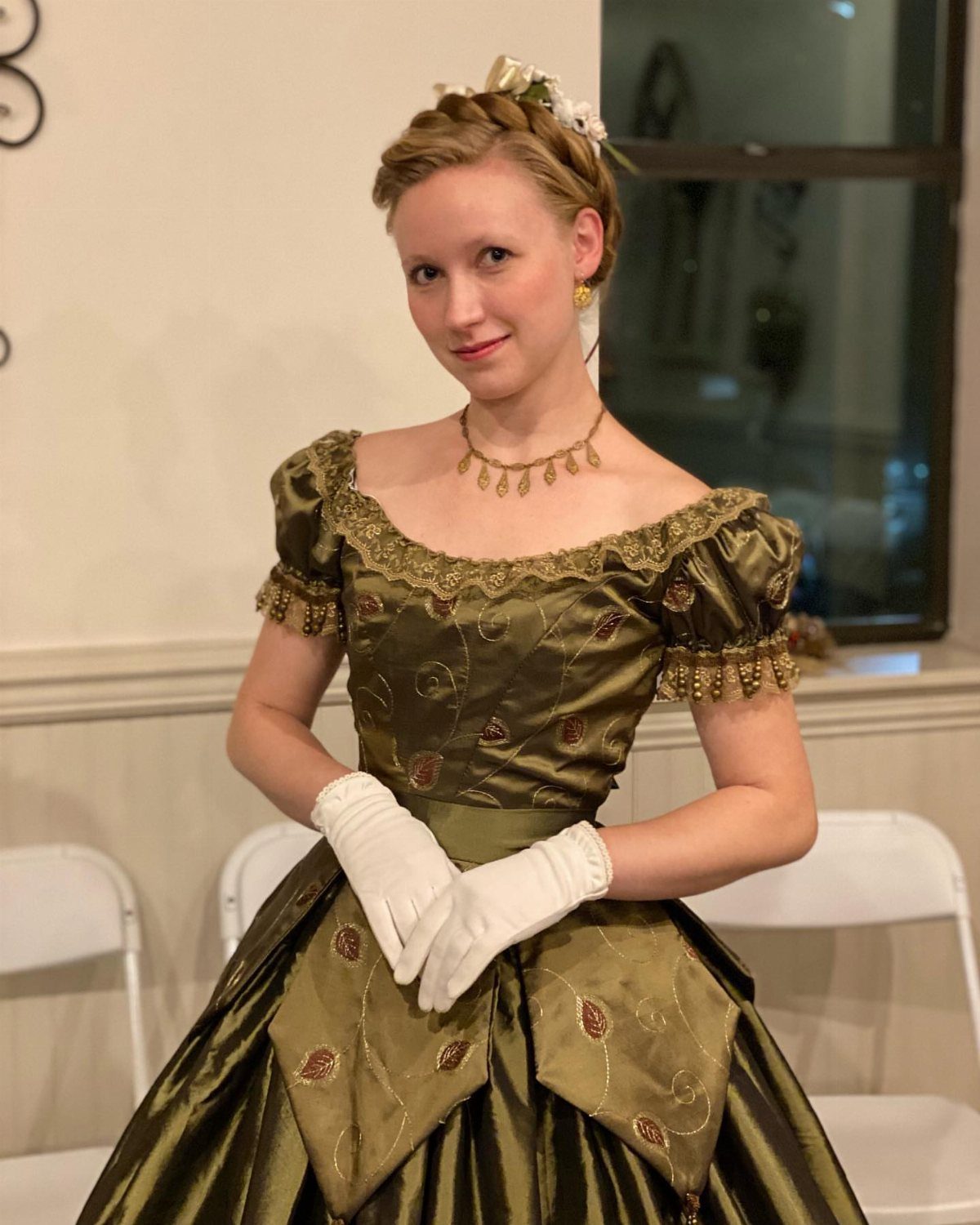 Woman in olive green satin Victorian gown with white gloves and gold jewelry.
