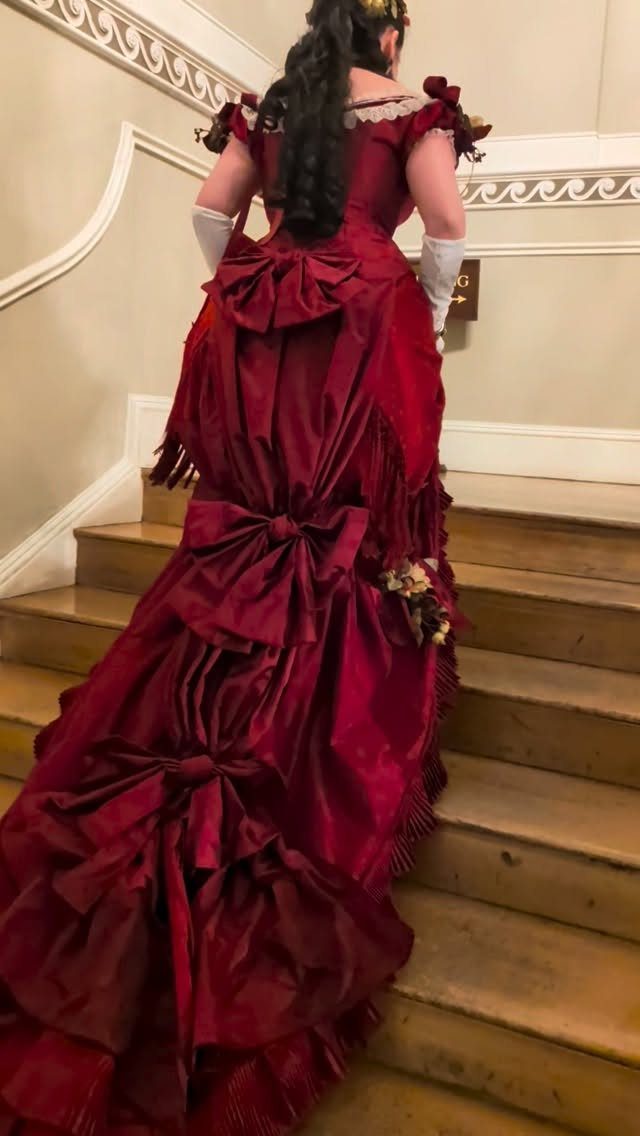 Woman in a red Victorian bustle gown with bows standing on wooden stairs.