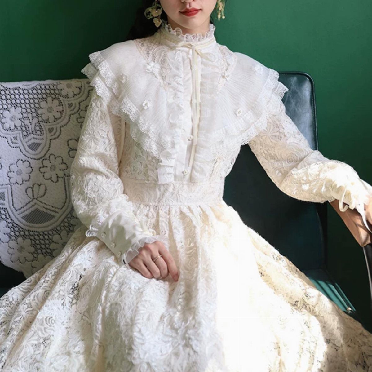 Woman wearing a high-collared white lace Victorian gown sitting against a green wall.