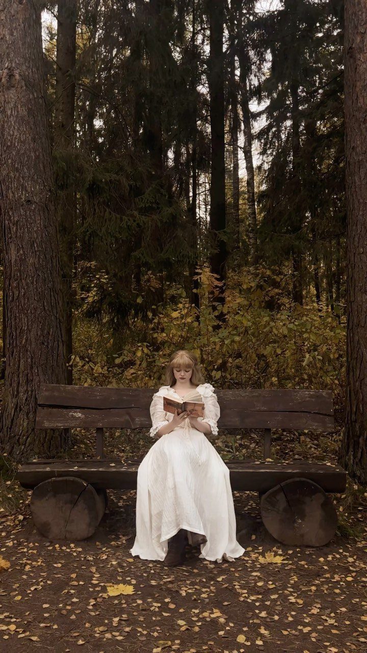 Woman in white Victorian gown reading a book on a forest bench